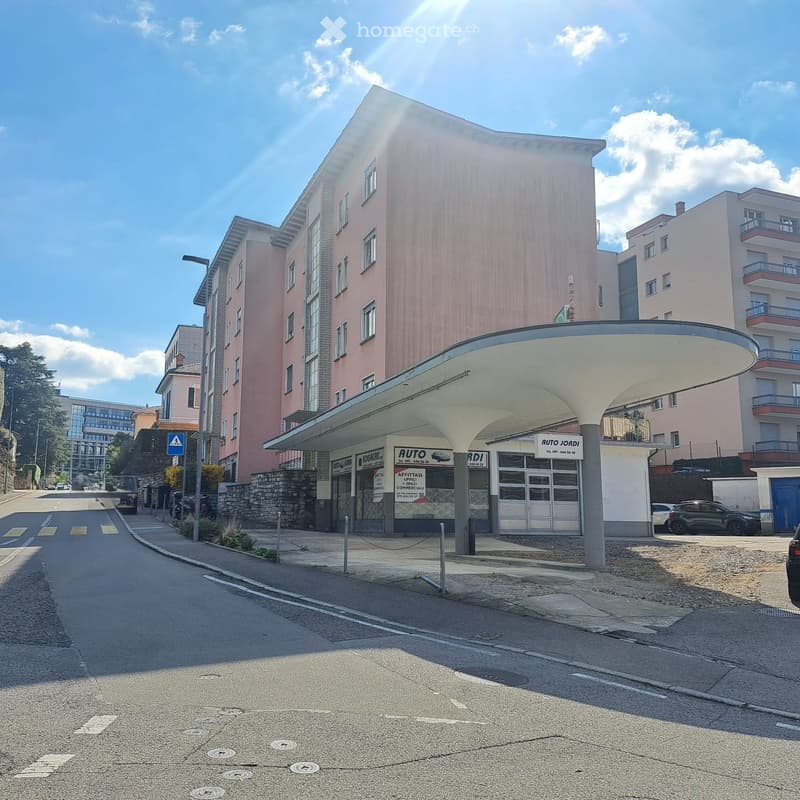 A building with a flat roof, multiple windows, and a sign that reads 'AUTO JARDI'. The building is situated on a street with parked cars and a sidewalk. There are trees and other buildings in the background.