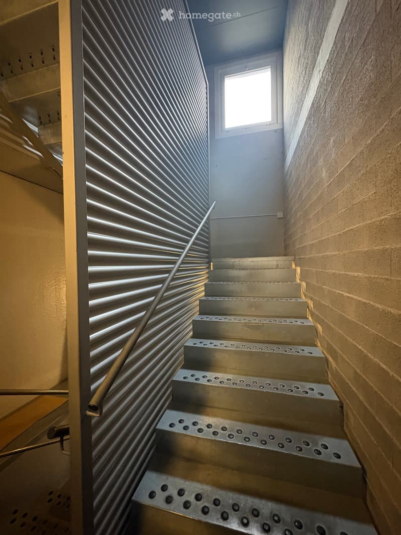 Stainless steel staircase with a metal handrail, located in a building with a brick wall and a window