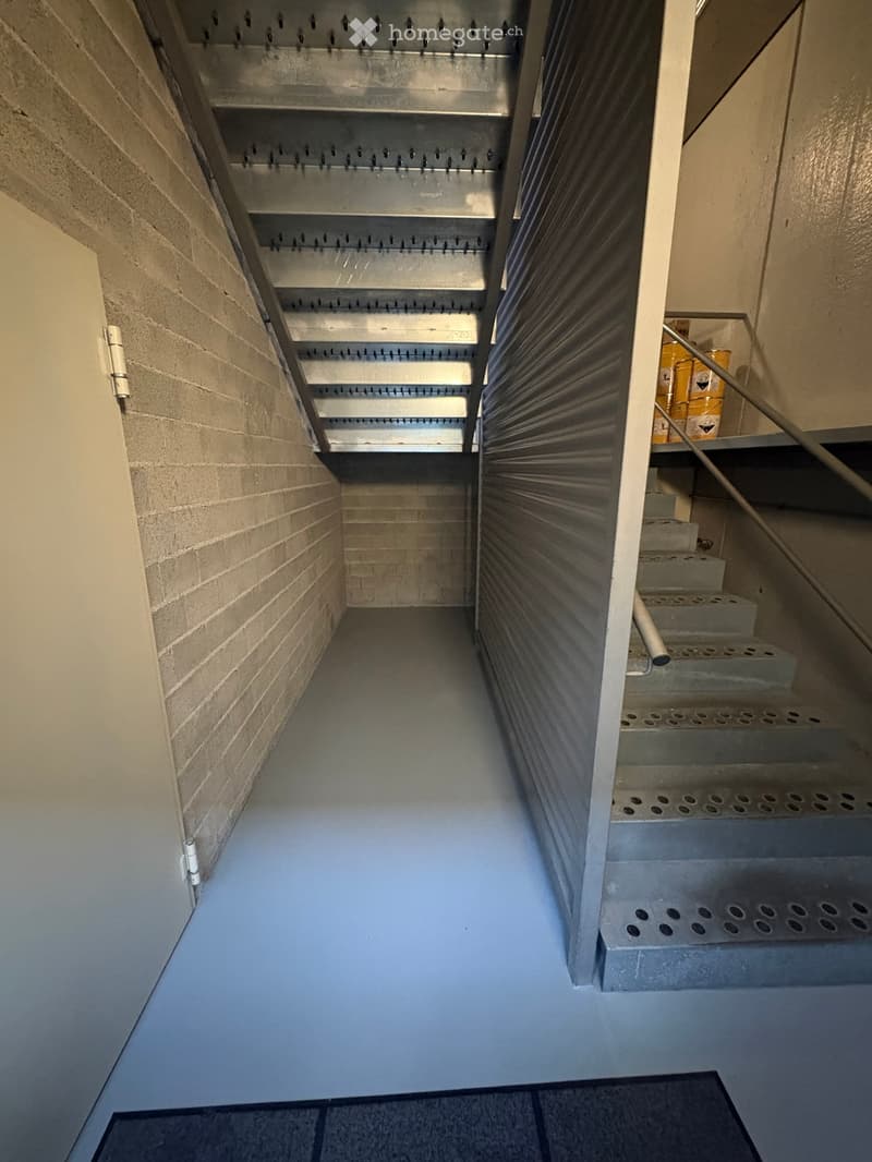 Concrete staircase with metal handrail, grey floor, brick wall, metal grates on the ceiling