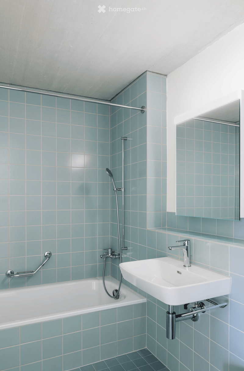 Modern bathroom with light blue tiled walls, a white bathtub, a white sink with a modern faucet, a showerhead, and a mirror above the sink.