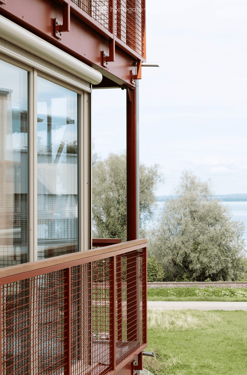 Balcony with red metal railing and glass window, overlooking a grassy area and water body.