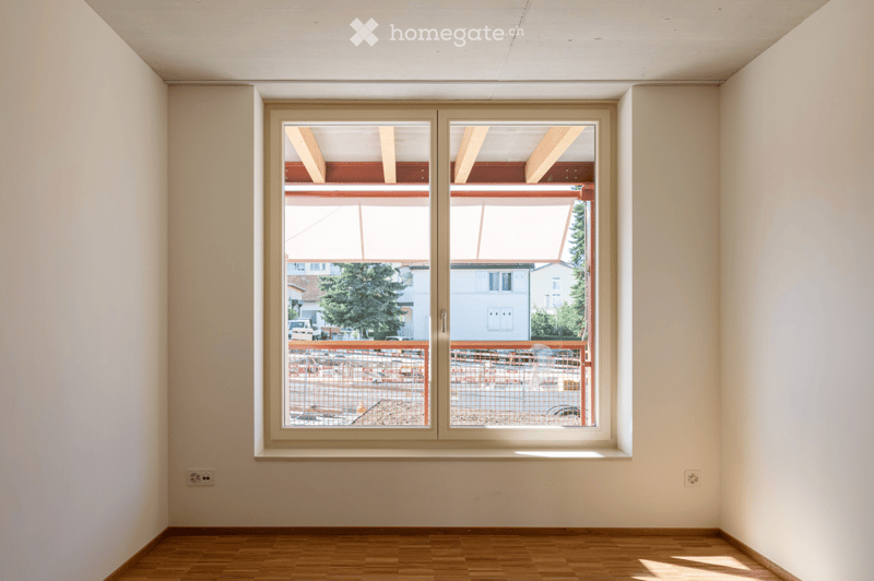Empty room with wooden flooring, white walls, and a large window offering a view of a construction site and surrounding buildings.