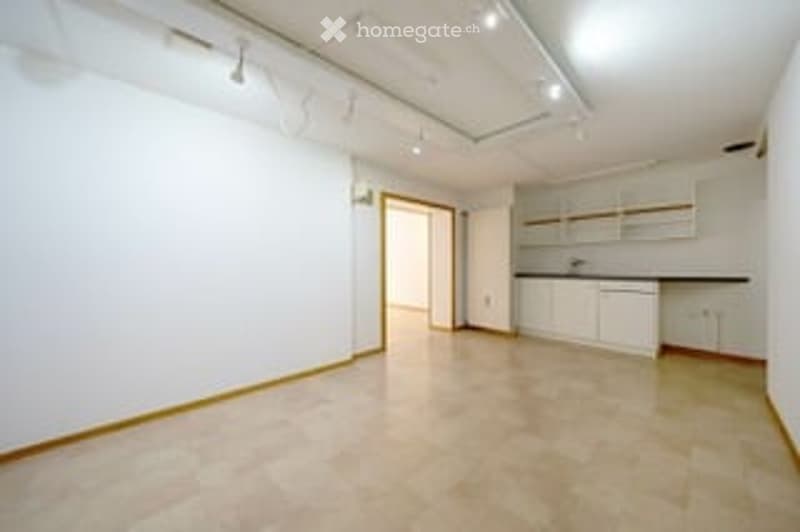 Empty room with white walls, beige tiles, open door, kitchen area with white cabinets, ceiling lights