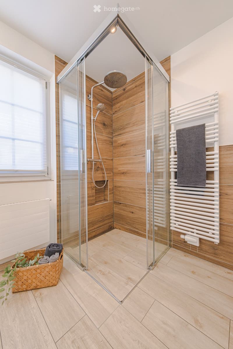 Modern bathroom with glass shower enclosure, wooden walls, tiled floor, and a towel rack with a towel