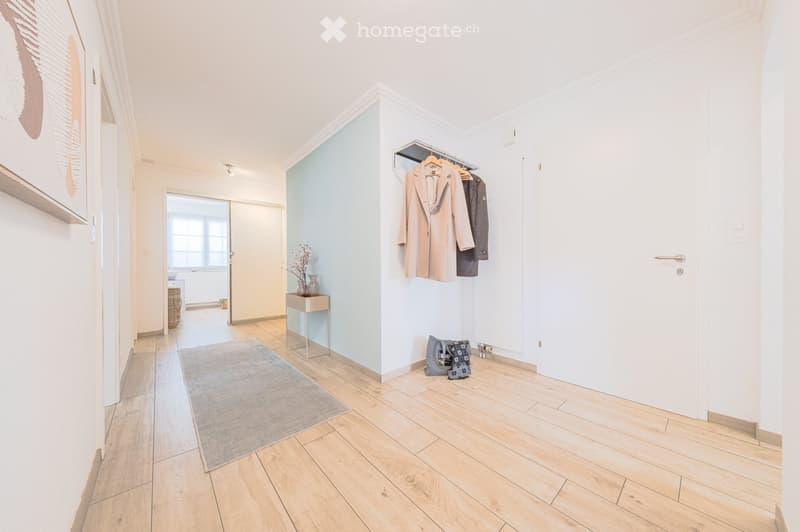 Empty hallway with wooden floor, white walls, blue accent wall, rug, coat rack, and a small table with a flower vase.