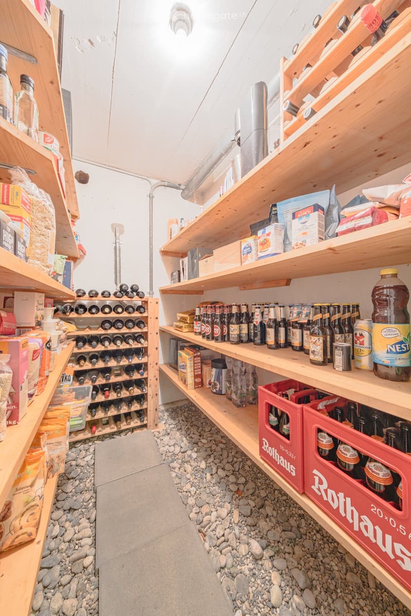 A storage room with wooden shelves, filled with various items such as bottles, boxes, and bags. The room has a gravel floor and a ceiling with a light fixture.