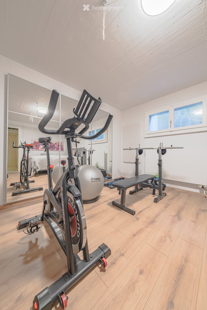 Home gym with exercise bikes, weight bench, and free weights. Large mirror on the wall. Wooden floor.
