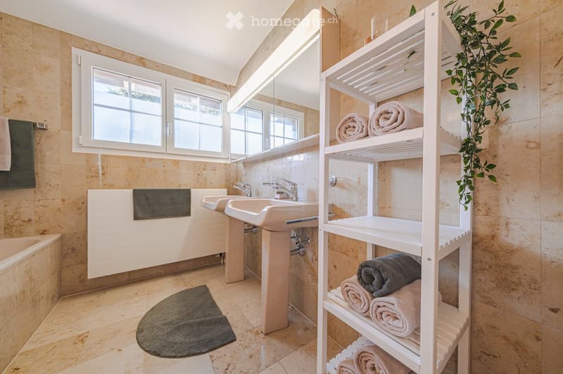 Bathroom with beige tiles, double sink, bathtub, towel rack, and a white shelf with towels.