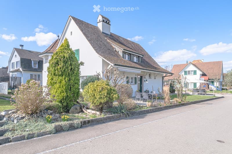 2 story house, white exterior, brown roof, green shutters, garden with plants and rocks, patio with table and chairs
