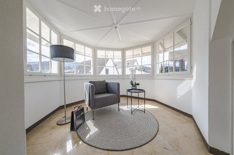 A room with a round rug, a gray armchair, a small table with a flower vase, a floor lamp, and large windows with a view of the outside.