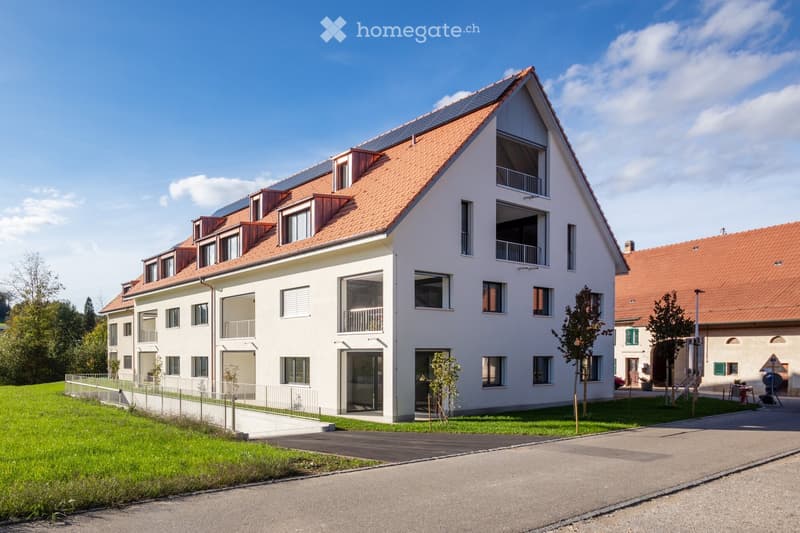 Modern white building with red roof, solar panels, multiple balconies, fenced yard, driveway