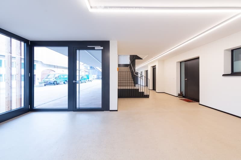 Empty room with white walls, beige floor, large glass doors, staircase, windows, and a view of the outside with cars.