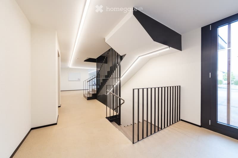 Empty room with white walls, beige floor, black railings, and a staircase.