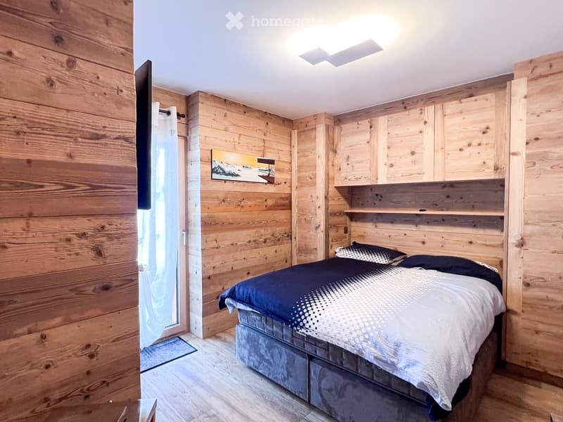 Bedroom with wooden walls, bed with blue and white bedding, TV mounted on wall, picture frame, sliding glass door, rug, ceiling light
