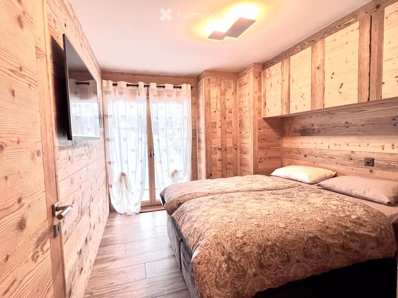 Bedroom with wooden walls, two beds, white curtains, TV mounted on wall, wooden floor, ceiling light