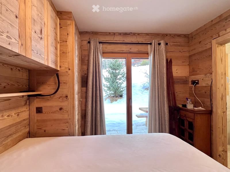 bedroom with wooden walls, large window with curtains, bed, wooden cabinet, wooden shelves, power outlet, view of snowy landscape