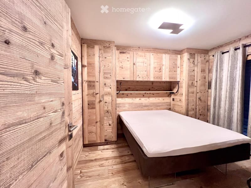 A small bedroom with wooden walls, a bed, and a shelf above the bed.