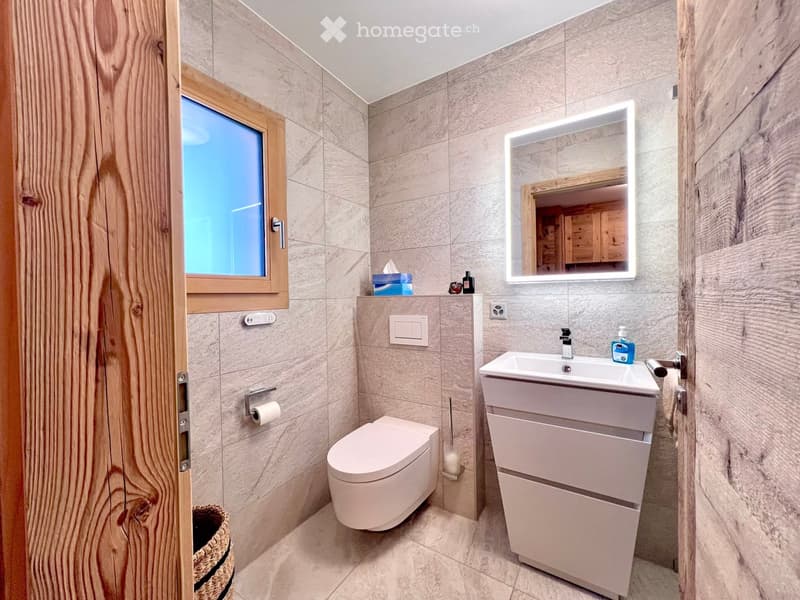 modern bathroom with white tiles, wooden walls, white toilet, white sink, mirror, window, toilet paper holder, basket, and toiletries