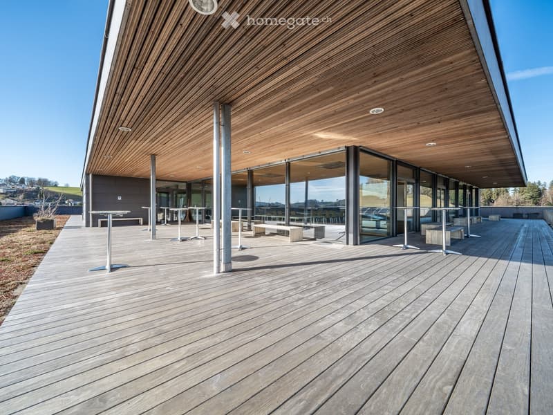 modern building with wooden deck, large glass windows, metal poles, tables and benches
