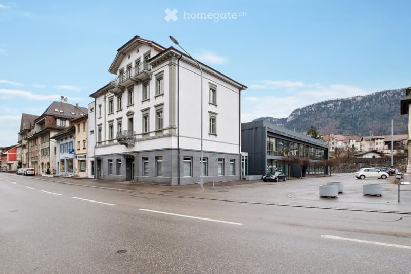 A three-story building with a white facade and gray accents, situated on a street with parked cars and streetlights. The building has multiple windows and balconies, and there are other buildings and a mountain in the background.