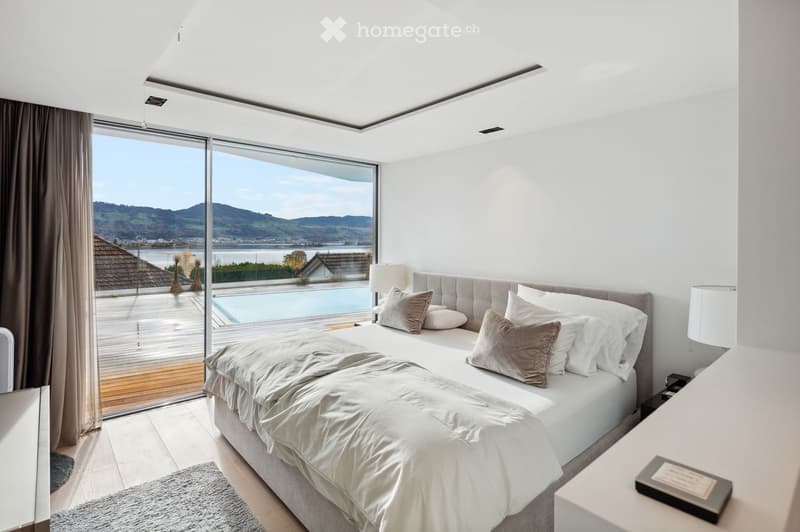 modern bedroom with large bed, white walls, wooden floor, large windows, balcony, view of lake and mountains
