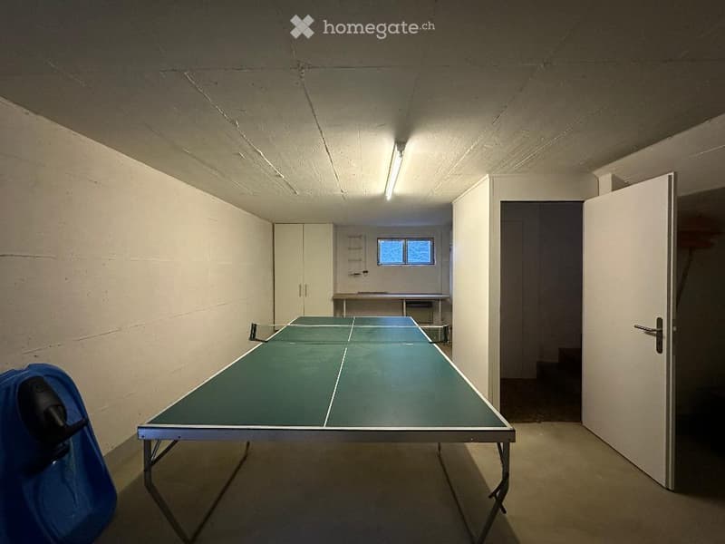 A room with a ping pong table, a blue chair, a white cabinet, a window, and a door.