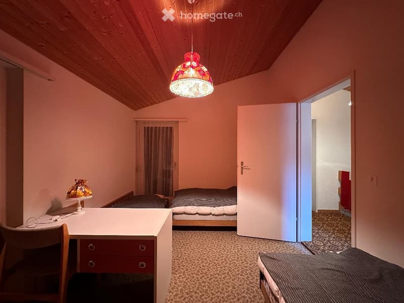 Bedroom with two single beds, desk, chair, lamp, wooden ceiling, carpeted floor, open door leading to another room