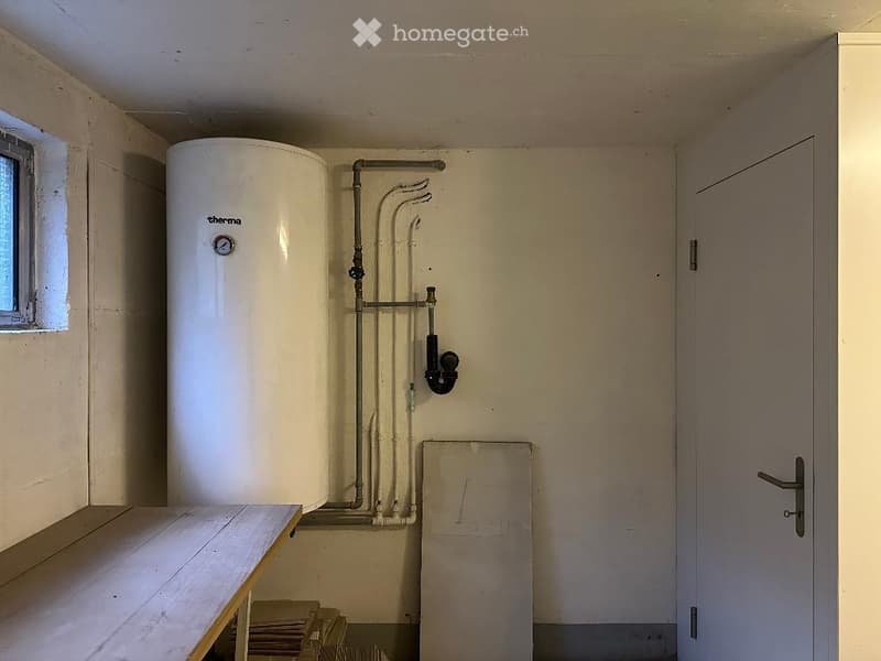 A room with a white water heater, pipes, a wooden table, and a window.