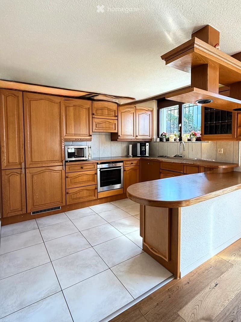 Wooden cabinets, stainless steel appliances, white tile flooring, breakfast counter, built-in microwave, coffee maker, and window with potted plants
