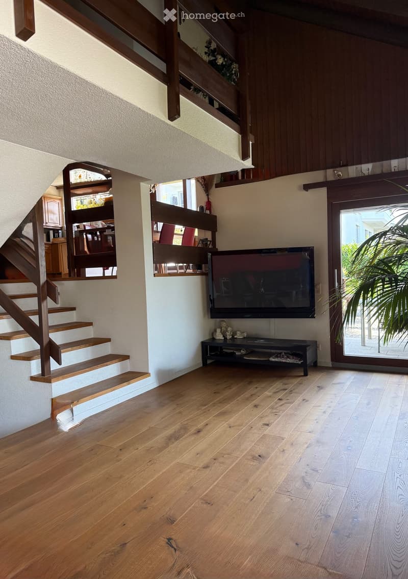 Open plan living room with a large TV, sofa, glass door to the balcony, wooden floor, and stairs