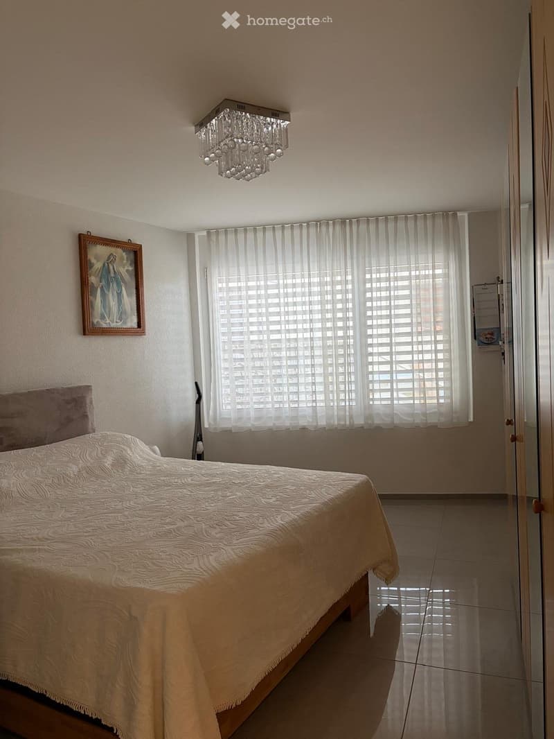 Bedroom with a bed, a chandelier, a picture frame, a window with white curtains, and a wooden floor.