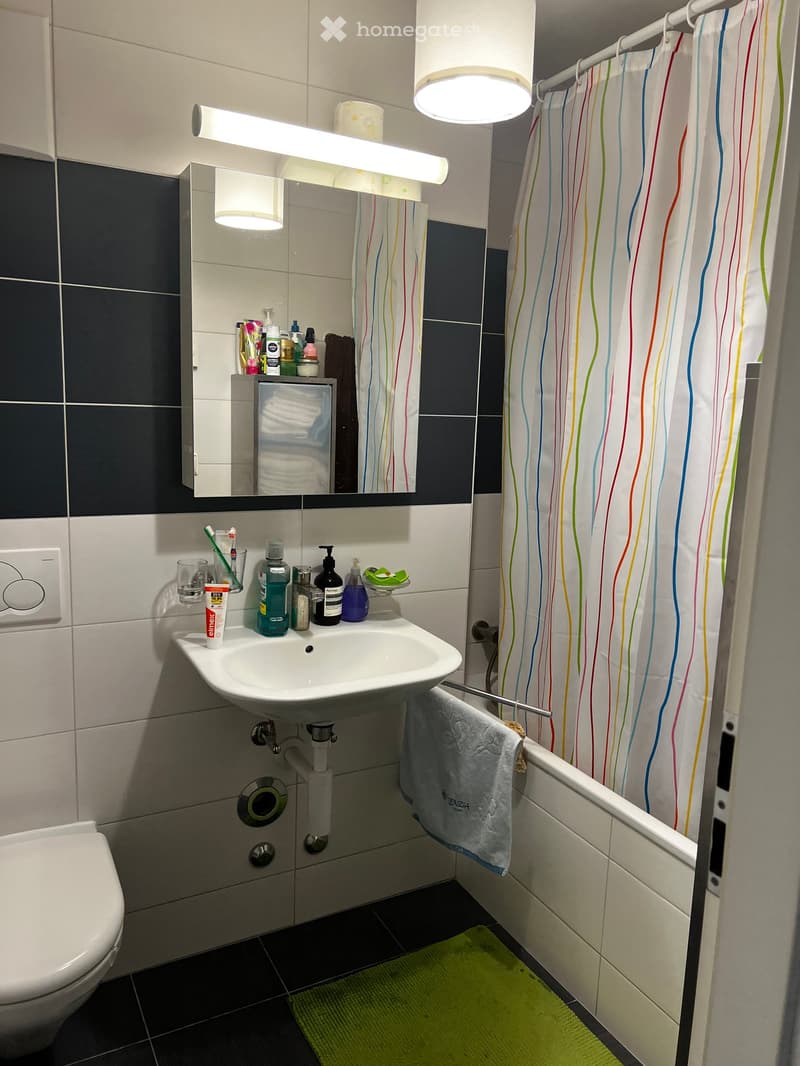 Bathroom with white tiles, sink, mirror, shower curtain, toilet, towel, and various toiletries