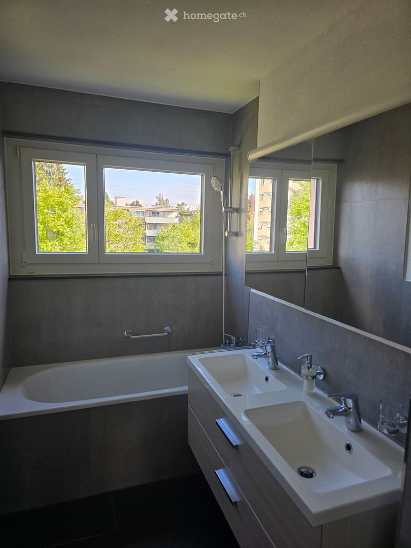 Modern bathroom with a bathtub, double sink, large mirror, and shower. Windows provide natural light and a view of greenery.