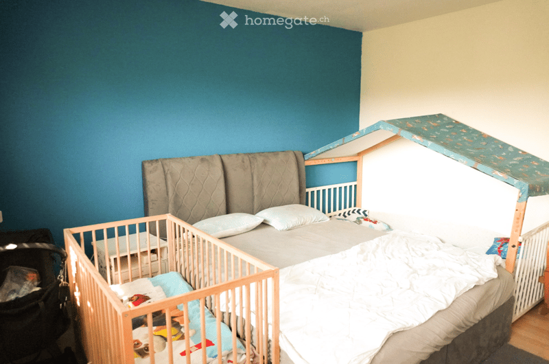 Bedroom with a double bed, crib, and a small cabinet. Blue walls, white ceiling, and wooden floor. There are toys on the floor.