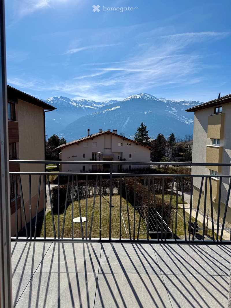 Balcony with metal railings, paved floor, view of a grassy area and a building with mountains in the background