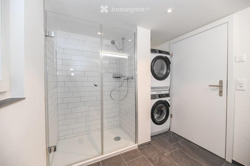 A small room with a glass shower, white tiled walls, a white washing machine, and a white dryer.