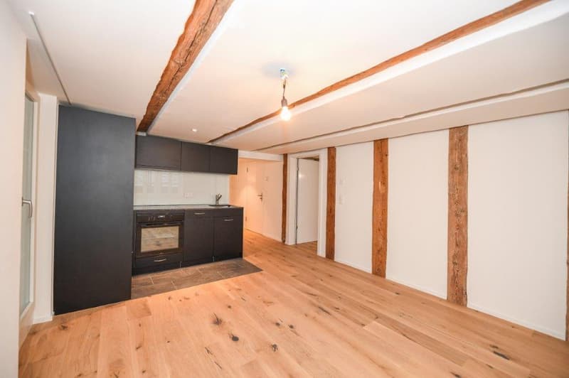 empty room, wooden floor, white walls, wooden beams, kitchen area with oven, cabinets and sink