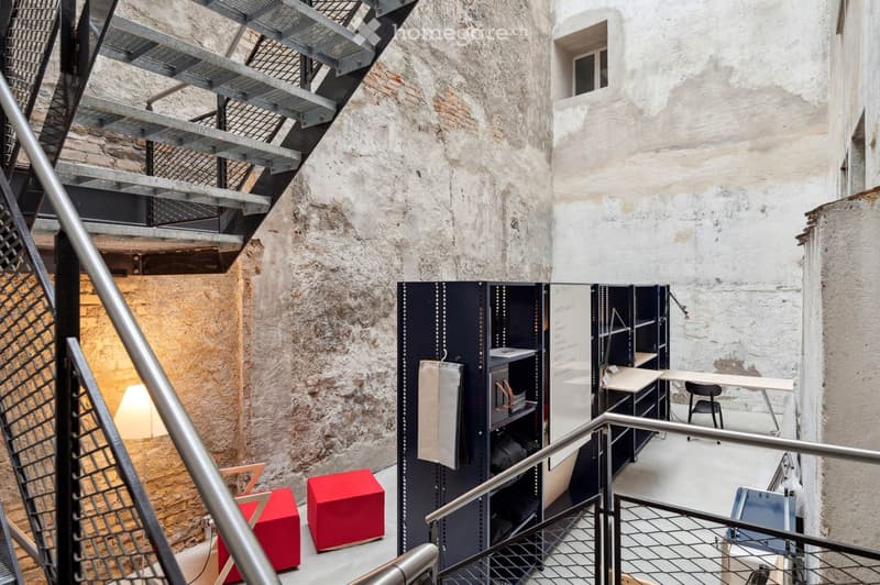 A room with exposed brick walls, metal staircase, metal racks with shelves, red stools, and an area with a desk and chair.