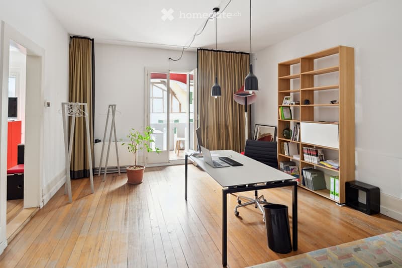 Wooden floor, white walls, desk with laptop, office chair, bookshelf, hanging lamps, radiator, open door, balcony