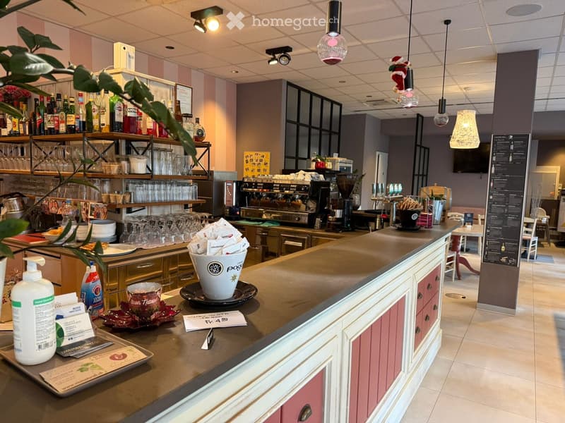 Counter with coffee machine, glasses, plates, and other items. Shelves with bottles and glasses. Tables and chairs in the background.