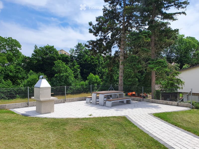 Outdoor seating area with a table and benches, a barbecue grill, and a paved pathway surrounded by grass and trees.