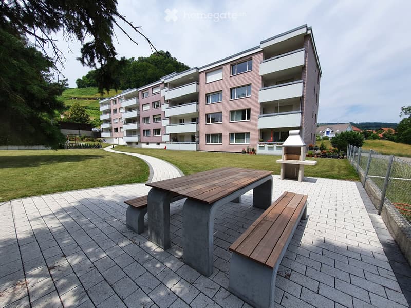 Outdoor seating area with a wooden table and benches, surrounded by a brick walkway, with a grassy area and a building in the background.