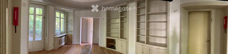 Empty room with wooden floor, white walls, large windows, built-in shelves, and a door.