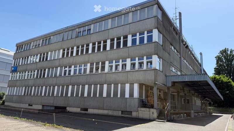 A multi-story concrete building with numerous windows and a flat roof, featuring a covered entrance area with stairs and railings.