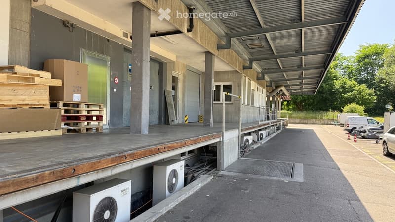 Warehouse or industrial building with a raised loading platform, stacked pallets, and air conditioning units visible.