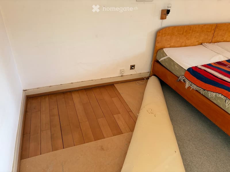 A small bedroom with a wooden bed, a colorful blanket, and a wooden floor. There is a large cardboard box in the foreground.