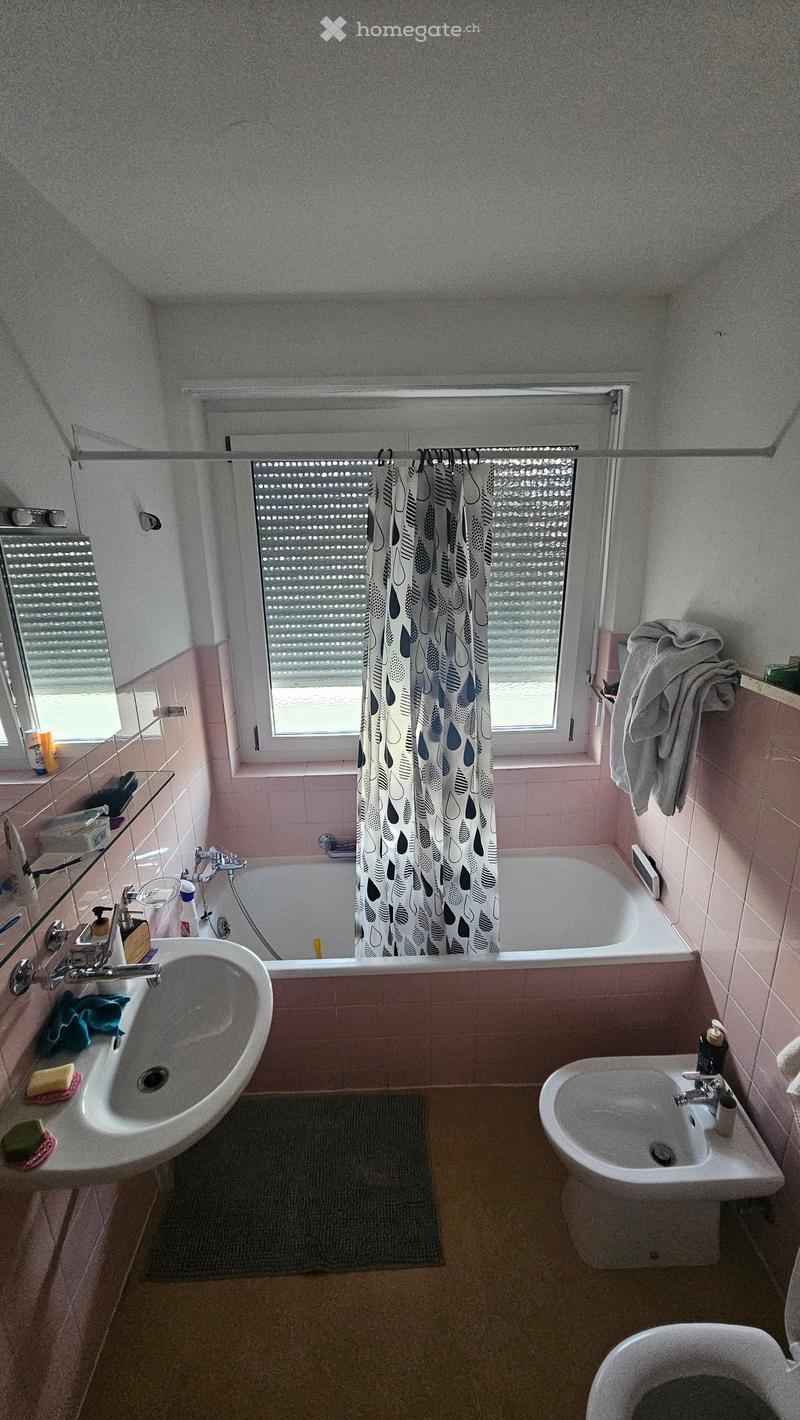 Bathroom with bathtub, sink, bidet, and toilet. Pink tiles, white fixtures, and a window with blinds.