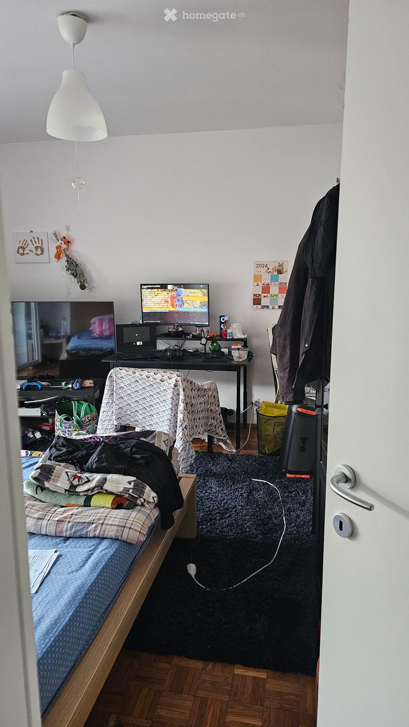 A bedroom with a bed, desk, monitors, and a chair. The room has a carpet, a lamp, and a calendar on the wall.