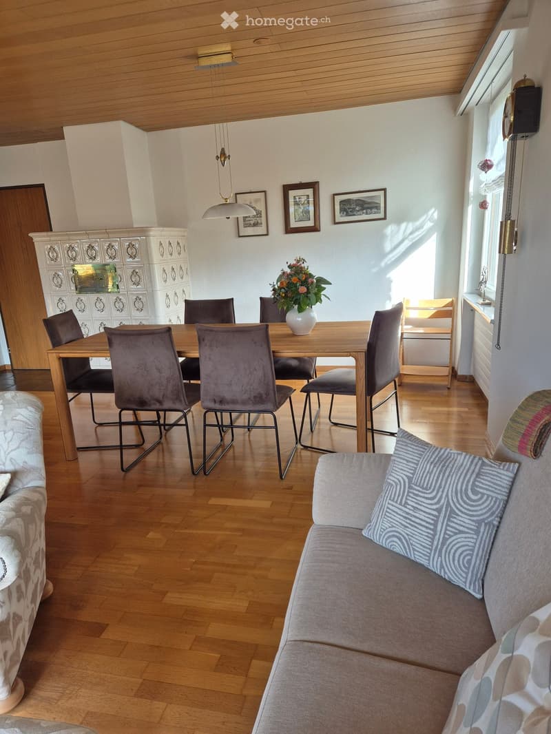 Dining room with wooden table, 6 chairs, flower vase, wooden cabinet, wooden floor, white walls, wooden ceiling, pendant light, framed pictures, clock, couch with pillows