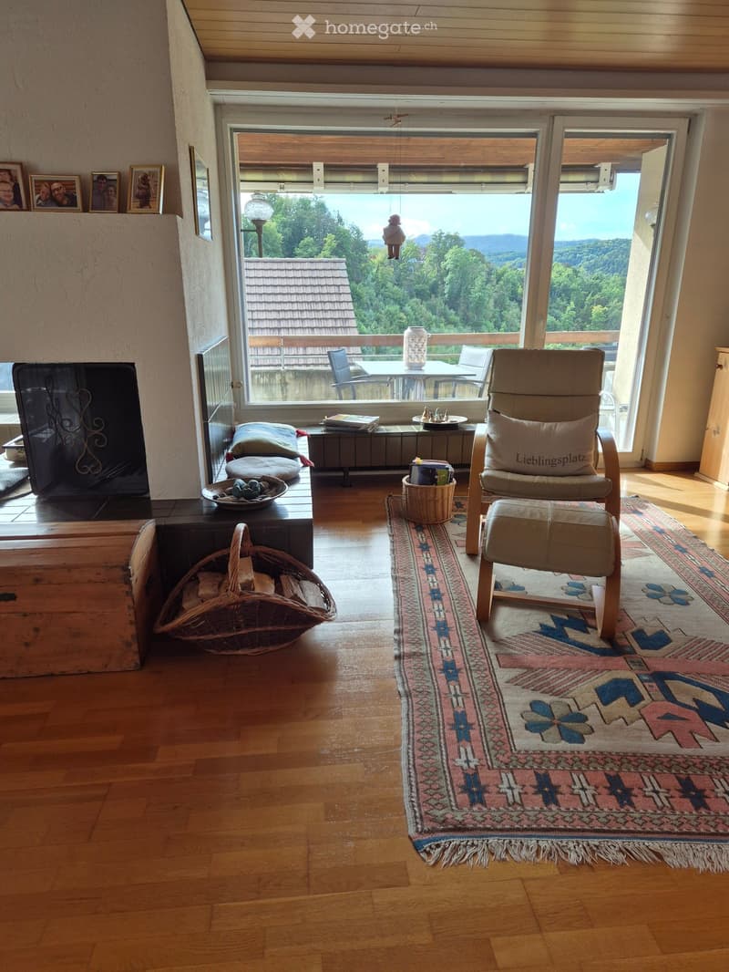 Living room with fireplace, large windows, wooden floor, armchair, rug, basket, storage box, table, chairs, and a view of the outside